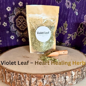 May include: A clear plastic bag filled with dried violet leaf herbs, labeled "Violet Leaf." A small wooden scoop and a pile of herbs sit on a wooden slice. The text "Violet Leaf - Heart Healing Herb" is at the bottom.