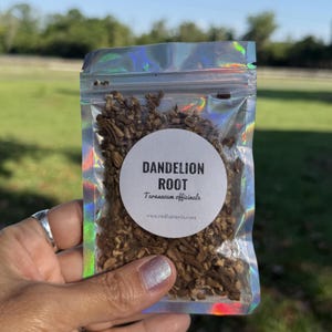 Puede incluir: Una bolsa holográfica transparente llena de raíz de diente de león seca. Una etiqueta circular blanca en la bolsa dice "Dandelion Root" con el nombre científico y una dirección de sitio web. La bolsa se sostiene en una mano.