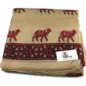 Puede incluir: Tela beige con un patrón repetido de bueyes rojos y un borde decorativo. Los bueyes están representados con sillas de montar y campanas. La tela está doblada, mostrando el patrón y la textura.