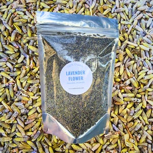 Può includere: Un sacchetto trasparente pieno di fiori di lavanda essiccati, etichettato "100% Organic Lavender Flower". Il sacchetto è circondato da boccioli di lavanda sfusi, che mostrano un'estetica naturale e terrosa. L'etichetta include il nome scientifico e un indirizzo web.