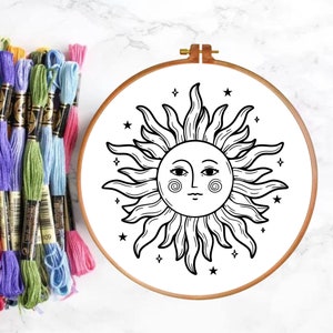 May include: A black and white embroidery hoop with a sun design. The sun has a face with eyes, a nose, and a mouth. There are small stars around the sun.