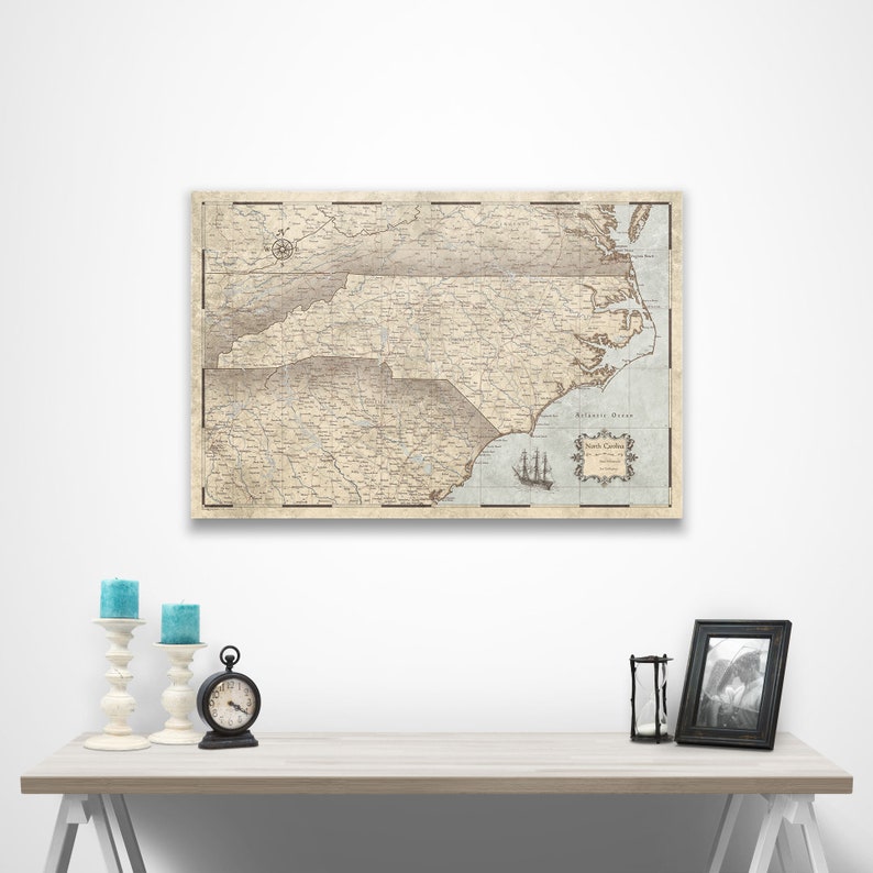 May include: A vintage-style map of North Carolina with a light blue background and brown lines. The map is framed and hanging on a white wall above a table with candlesticks and a clock.