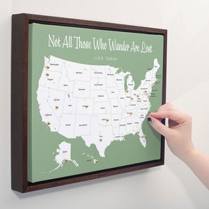 May include: A green and white map of the United States with gold push pins marking locations. The map is framed in a dark brown wood frame. The quote "Not All Those Who Wander Are Lost - J.R.R. Tolkien" is printed at the top of the map.
