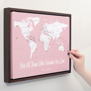 Peut inclure: Une carte du monde rose avec des punaises dorées, encadrée de bois foncé. La carte est sur un mur blanc. La citation "Not All Those Who Wander Are Lost - J.R.R. Tolkien" est imprimée sous la carte.