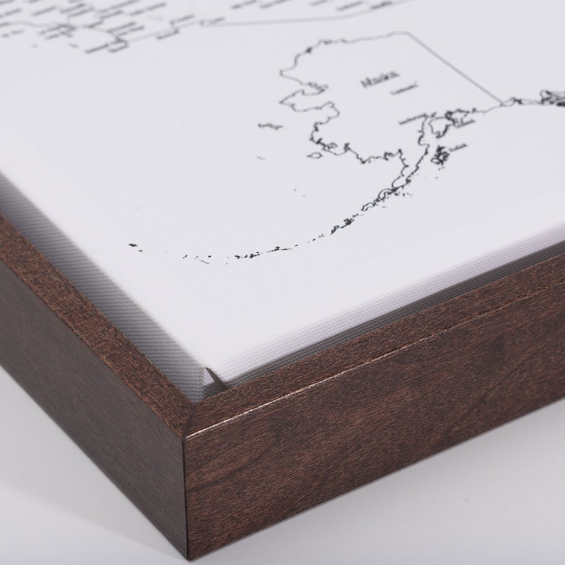 May include: A close-up of the corner of a framed canvas print of a map of Alaska. The frame is made of dark brown wood.
