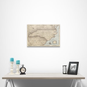 May include: A vintage-style map of North Carolina with a light blue background and brown lines. The map is framed and hanging on a white wall. There are candles and a clock on a white table in front of the map.
