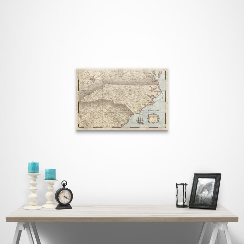 May include: A vintage-style map of North Carolina with a light blue background and brown lines. The map is framed and hanging on a white wall. There are candles and a clock on a white table in front of the map.