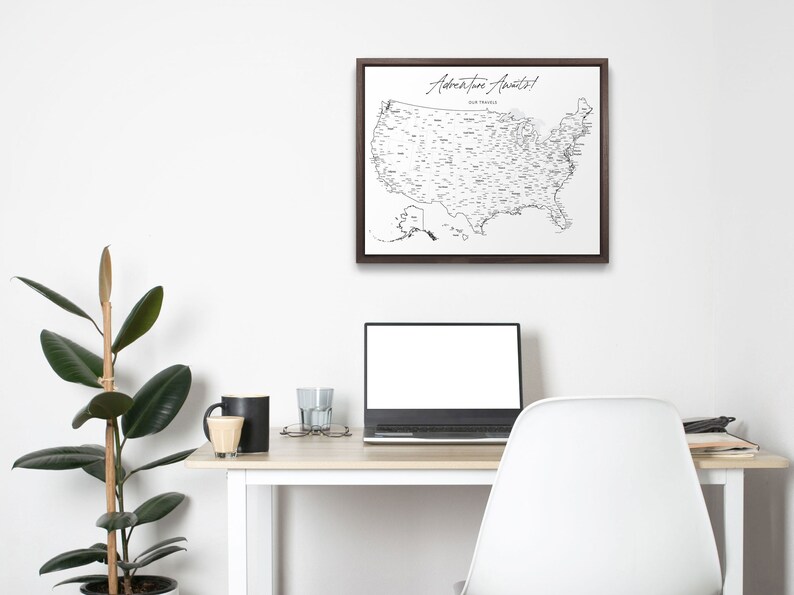 May include: A black and white map of the United States with the text "Adventure Awaits!" in a brown frame hanging on a white wall above a desk with a laptop, a plant, and a coffee mug.