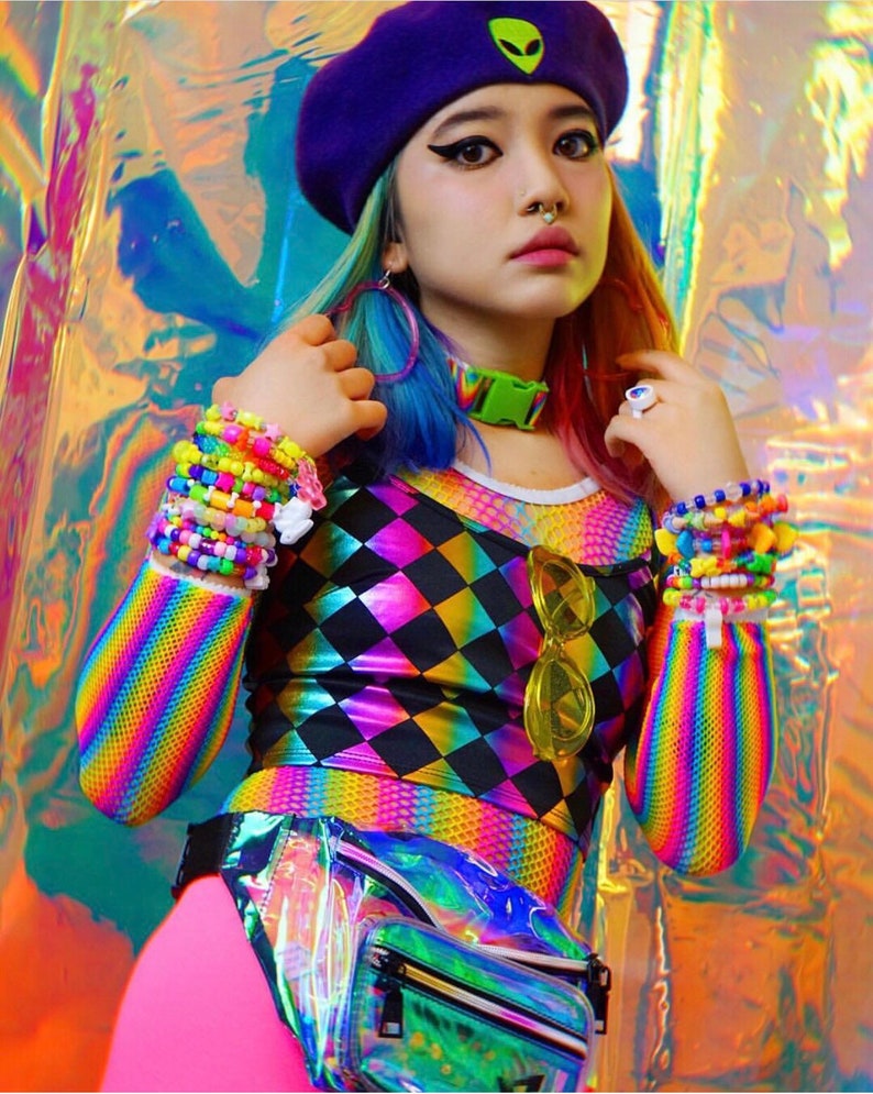 May include: A woman wearing a rainbow striped long-sleeved shirt with a black and rainbow diamond pattern top. She is wearing a purple beret with a green alien graphic, a rainbow beaded bracelet, and a clear iridescent fanny pack.