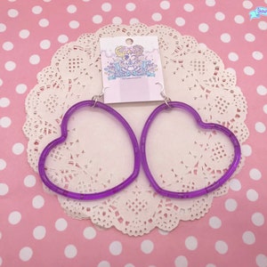 May include: A pair of purple heart-shaped hoop earrings. The earrings are made of clear plastic with a purple tint. The earrings are on a white doily with a pink polka dot background.