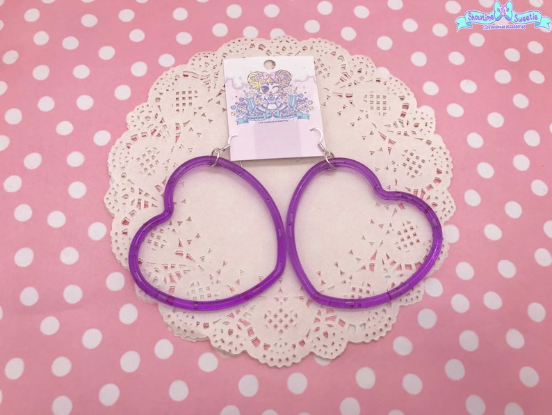 May include: A pair of purple heart-shaped hoop earrings. The earrings are made of clear plastic with a purple tint. The earrings are on a white doily with a pink polka dot background.