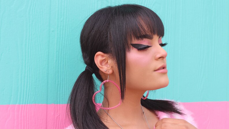 May include: A woman with black hair and bangs wearing pink heart-shaped earrings. She is wearing a white fur jacket and has dramatic black eyeliner.