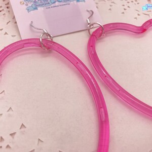 May include: A pair of pink heart-shaped hoop earrings with silver hooks. The earrings are made of a clear plastic material.