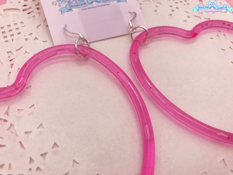 May include: A pair of pink heart-shaped hoop earrings with silver hooks. The earrings are made of a clear plastic material.