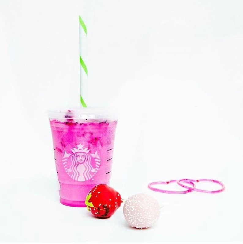 May include: A pink Starbucks cup with a green and white striped straw, filled with a pink drink.  There is a pink and white cake pop and a red and white cake pop next to the cup.