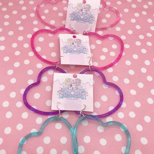 May include: Four pairs of heart-shaped hoop earrings in different colors. The earrings are made of clear plastic with a gradient of color. The colors are pink, purple, and blue.