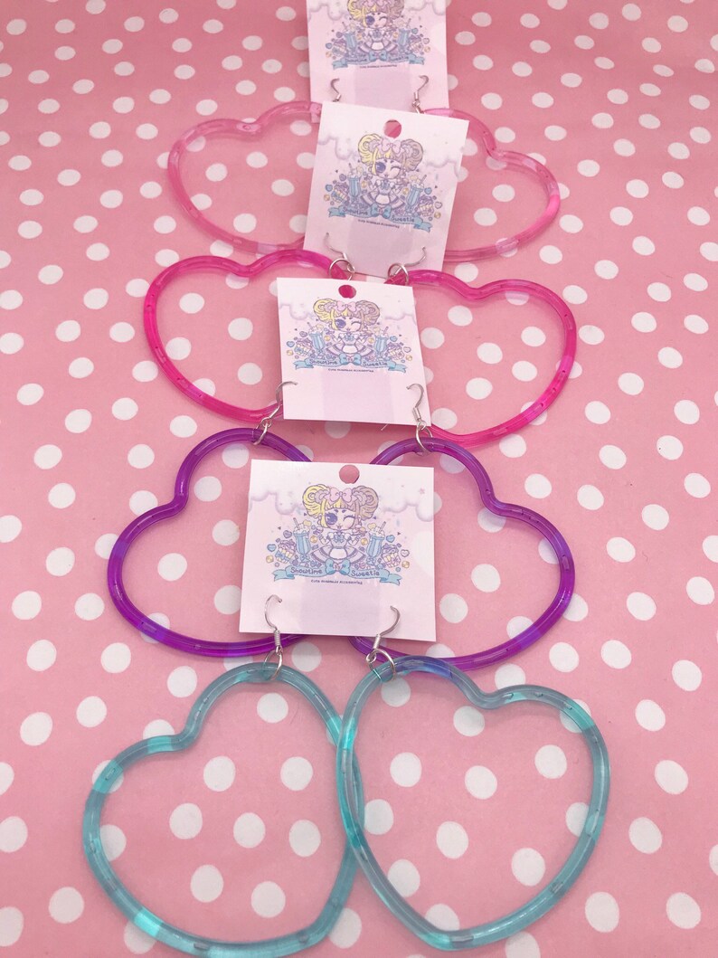 May include: Four pairs of heart-shaped hoop earrings in different colors. The earrings are made of clear plastic with a gradient of color. The colors are pink, purple, and blue.