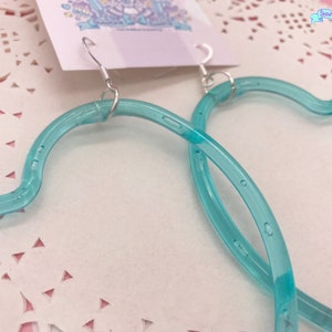 May include: A pair of turquoise blue heart-shaped hoop earrings. The earrings are made of a clear plastic material and have a shiny finish.