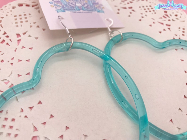 May include: A pair of turquoise blue heart-shaped hoop earrings. The earrings are made of a clear plastic material and have a shiny finish.