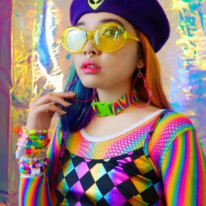 May include: A person wearing a purple beret with an alien face embroidered on it, yellow round sunglasses, a rainbow striped long-sleeved shirt, a black and white diamond patterned top, and a green buckle choker.