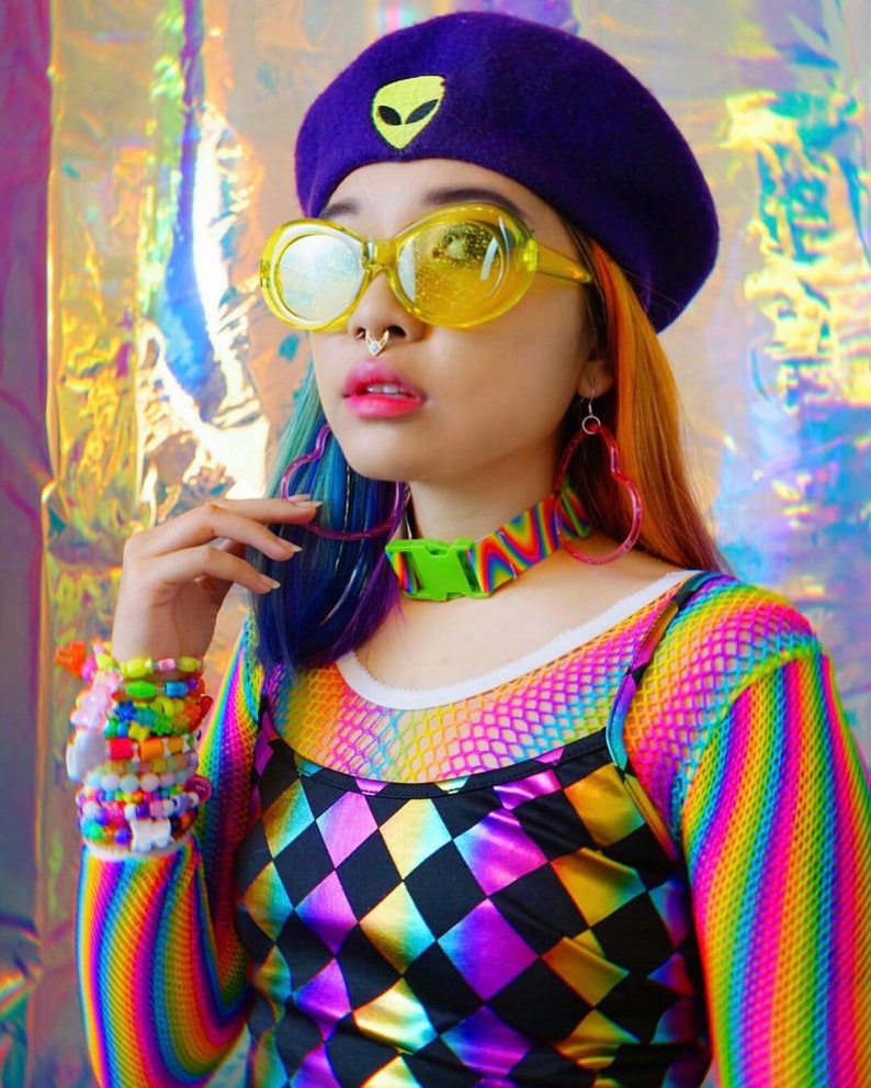 May include: A person wearing a purple beret with an alien face embroidered on it, yellow round sunglasses, a rainbow striped long-sleeved shirt, a black and white diamond patterned top, and a green buckle choker.