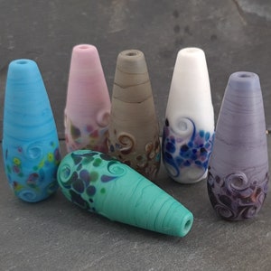 Peut inclure: Six perles en verre de différentes couleurs et motifs, notamment bleu, rose, gris, blanc et violet. Les perles sont en forme de goutte et présentent des tourbillons et des points.