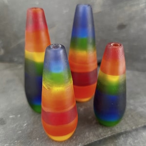 May include: Four glass rainbow-coloured teardrop-shaped ornaments. The ornaments are layered with different colours of glass, including red, orange, yellow, green, blue, and purple.