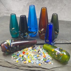 May include: A collection of colourful glass beads with a variety of patterns and designs. The beads are arranged on a dark grey surface with a pile of smaller glass pieces in the centre.