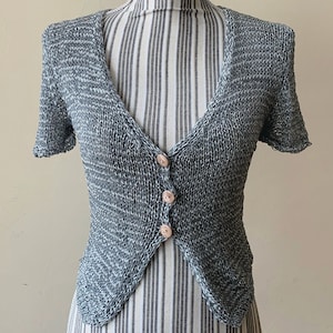 Puede incluir: Un cárdigan de punto gris y blanco con cuello en V y mangas cortas. El cárdigan tiene tres botones en la parte delantera.