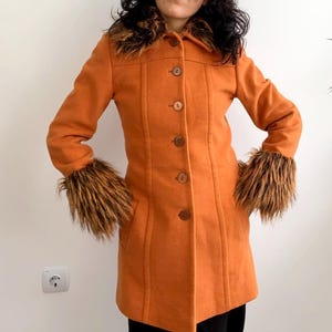 May include: Orange coat with brown fur trim on the collar and cuffs. The coat has a button-up front and two front pockets. The woman is wearing black pants. The coat is a vintage style.