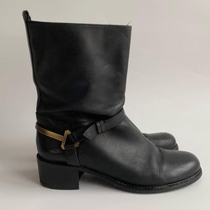 May include: A black leather mid-calf boot featuring a rounded toe, a low block heel, and a decorative strap across the ankle secured with a metallic gold-toned hardware accent. The boot has a pull tab at the top rear and a smooth finish.