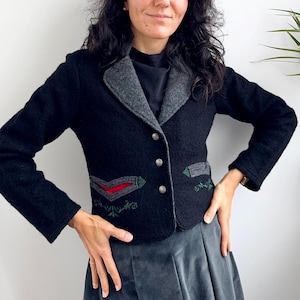 May include: A black wool jacket with a grey collar and decorative embroidery on the pockets. The jacket has three silver buttons and a tailored fit. The woman is wearing a grey skirt.