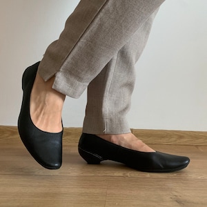 May include: A pair of black leather heeled shoes with a pointed toe. The shoes are worn by a person wearing beige pants.