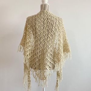 Könnte beinhalten: Ein cremefarbener Häkel-Schal mit Fransen. Der Schal wird auf einer weißen Schaufensterpuppe präsentiert.