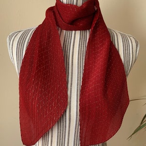 May include: A red pleated scarf with gold thread accents. The scarf is displayed on a white mannequin with a striped torso.