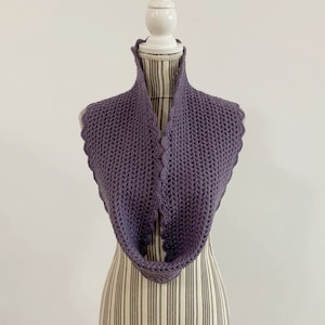 以下が含まれることがあります： 白いマネキンに飾られた紫色のクロシェのネックウォーマー。ネックウォーマーは、波打つ縁取りと、質感のあるステッチでできています。