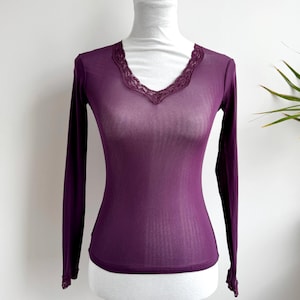 May include: A long-sleeved, sheer plum-colored top with a V-neckline. The neckline is trimmed with delicate lace. The top is displayed on a white mannequin against a white background.