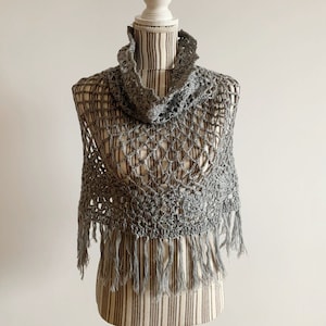 May include: A gray crocheted shawl with a fringe border. The shawl is displayed on a white mannequin.