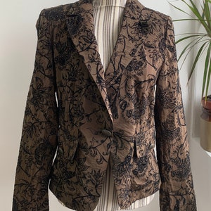 May include: A brown blazer with a black floral pattern. The blazer has a single button fastening and a pocket on the left side.