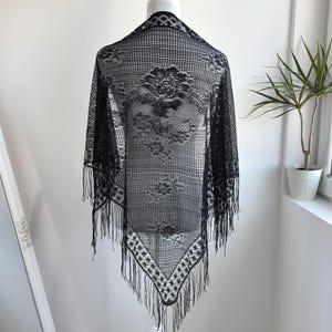 May include: A black lace shawl with a floral pattern and fringe trim. The shawl is displayed on a white mannequin, showcasing its intricate design and delicate texture. The shawl is a triangular shape.