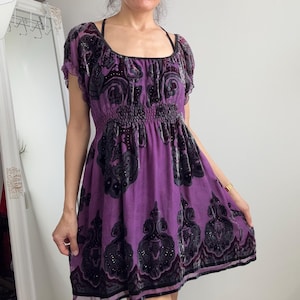 May include: A short, purple dress with a scoop neckline and short sleeves. The dress features a black paisley pattern and a gathered waist. The fabric has a slight sheen. The dress is knee-length.