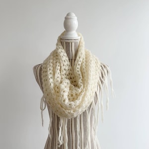 May include: A white crocheted scarf with fringe on a white mannequin. The scarf is a light cream color and has a delicate openwork pattern.
