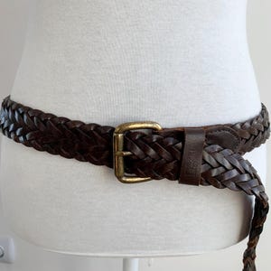 May include: A dark brown, plaited leather belt with a gold-coloured buckle. The belt is displayed on a white mannequin. The woven design of the belt adds texture and style. A classic accessory for any wardrobe.