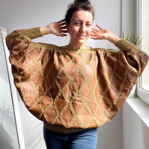May include: A brown leather top with a diamond pattern and gold stitching. The top has long sleeves and a wide neckline. The model is wearing blue jeans.