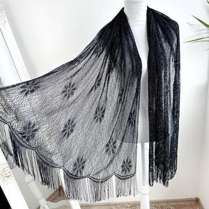 May include: A black lace shawl with a floral pattern and fringe trim. The shawl is draped over a white mannequin, showcasing its delicate design and texture. The background is a plain white wall.