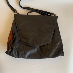 Könnte beinhalten: Schwarze Leder-Umhängetasche mit Klappenverschluss und brauner Seitenwand. Die Tasche hat einen langen, verstellbaren Schulterriemen und einen oberen Griff. Die Tasche ist ca. 36 cm breit und 41 cm hoch.