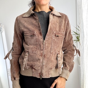 Könnte beinhalten: Braune Cordjacke mit Reißverschluss, zwei Brusttaschen und beigen gerippten Bündchen und Saum. Die Jacke hat einen Kragen und Riemen an den Ärmeln. Getragen über einem grauen Oberteil und kombiniert mit einem schwarzen Rock.