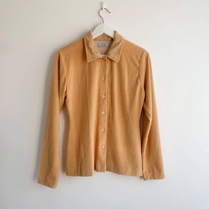 May include: A long-sleeved, button-up shirt in a light orange colour. The shirt has a collar and a small label that says "Mille" on the front.
