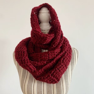 以下が含まれることがあります： 濃い赤色のクロシェの無限スカーフ。スカーフは太い糸で作られており、シンプルで質感のあるステッチが施されています。