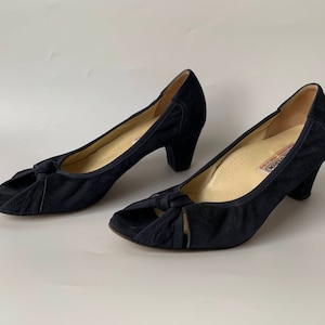 May include: A pair of navy blue suede peep-toe heels. Each shoe has a knotted detail on the vamp and a small, rounded heel. The interior of the shoe is a light beige colour.